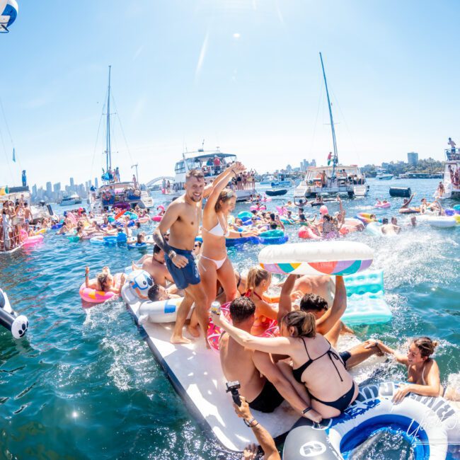 A lively scene of people having fun in the water near several boats. Many are on colorful inflatable floats, enjoying the sunny day with skyscrapers visible in the distance. The atmosphere is festive with many people gathered on a floating platform.