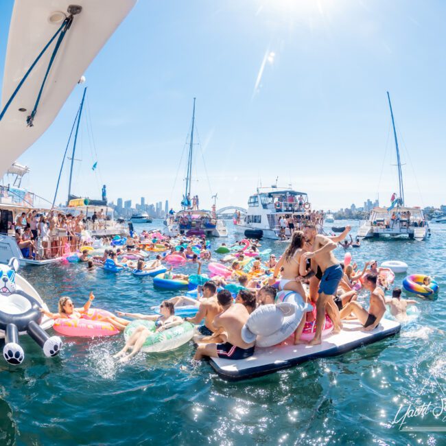 A festive pool party on the water with people on inflatables and floats, surrounded by boats under a sunny blue sky. The scene is lively with various colorful inflatables and people enjoying the event.