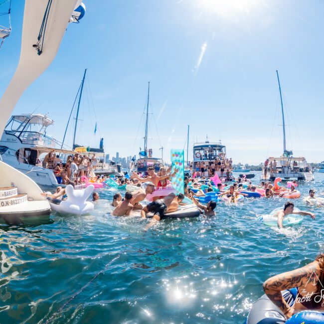 A lively boat party on a sunny day with people on colorful inflatables in the water. Several boats are anchored nearby, and the skyline is visible in the distance under a clear blue sky.