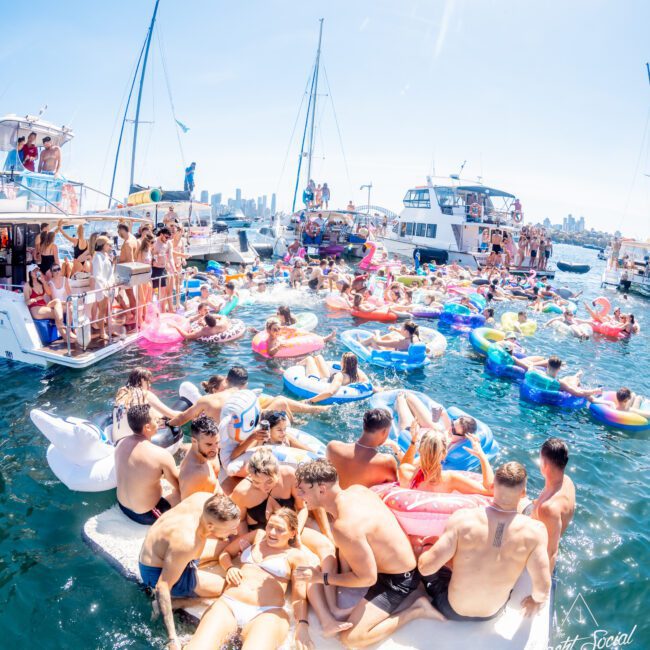 A lively boat party on a sunny day with many people on inflatable floats in the water. Several yachts are anchored nearby as attendees enjoy the vibrant atmosphere. The city skyline is visible in the background under a clear blue sky.