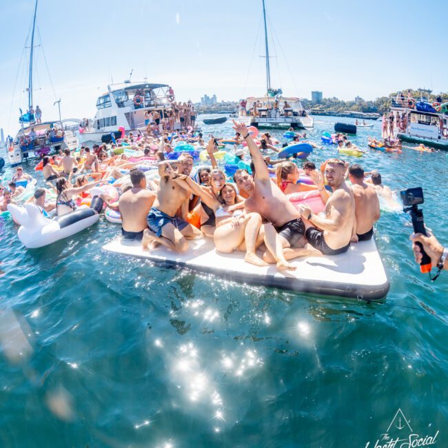 A lively scene on the water with a group of people enjoying a boat party. They are sitting on a large inflatable near yachts, surrounded by colorful floats under a bright blue sky. The atmosphere is festive and joyful.