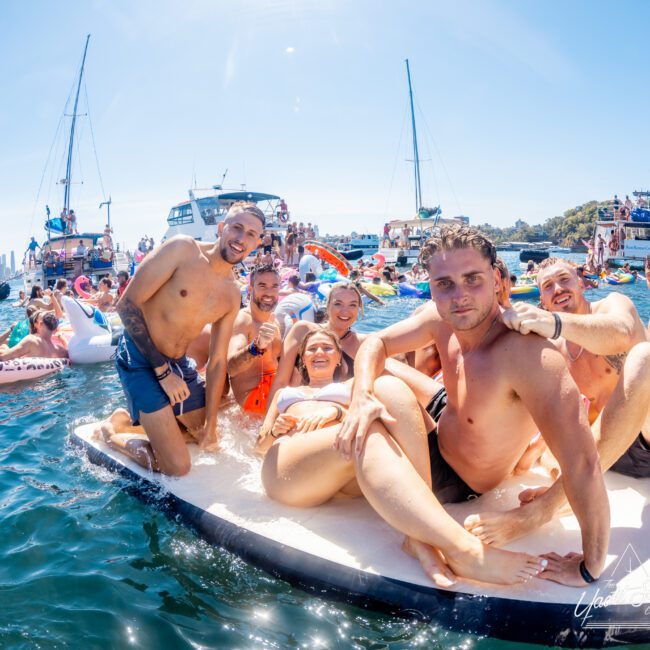 A group of people in swimwear are relaxing and smiling on a floating platform in a busy, sunlit harbor. Surrounding them are boats and colorful inflatables, with a city skyline visible in the background.