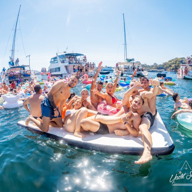 A group of people on a floating platform enjoy a sunny day on the water, surrounded by boats and inflatables. The city skyline is visible in the background, and the people are smiling and laughing.