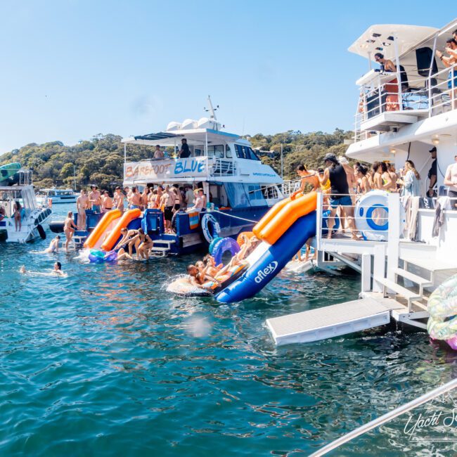 A lively scene with people enjoying a boat party. Two boats are docked, and guests are swimming and using inflatable slides. The day is sunny, and the water is a clear blue. Trees are visible in the background.