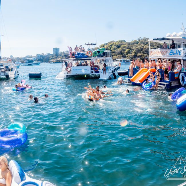 People are enjoying a sunny day on a vibrant blue waterfront, with several boats and inflatables. Some individuals are swimming, while others socialize on the boats. A slide extends from one boat into the water, adding to the lively scene.