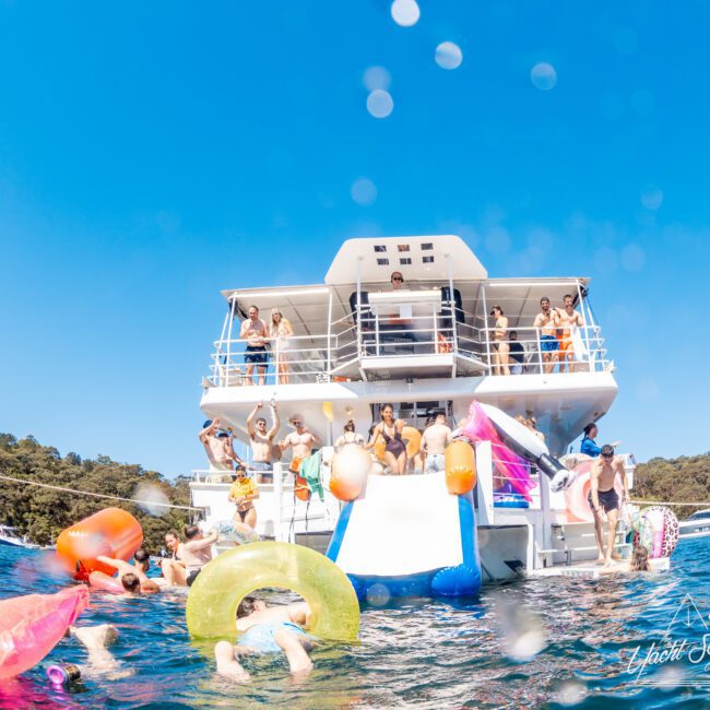A group of people enjoying a sunny day on a yacht. Some are on the deck and others are in the water with colorful floaties, including a unicorn. The sky is clear and blue, and the scene is lively and festive.