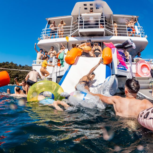 A vibrant scene of people enjoying a sunny day on a boat with inflatable toys. Some are sliding into the water via a slide at the back of the boat. The background features green hills under a clear blue sky.