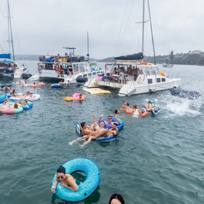 A lively scene unfolds as a Social Club gathers on the water, with people partying on inflatable tubes and boats. Several yachts are anchored close together, their occupants socializing and relaxing in the cloudy ambiance against a forested backdrop.