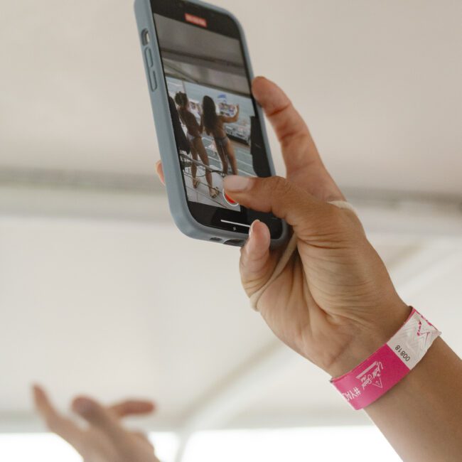 A person holds a smartphone, capturing a photo of people in swimwear by a pool. They wear a pink wristband, and the phone's screen shows the image being taken. The background is blurred, suggesting a casual, lively atmosphere.