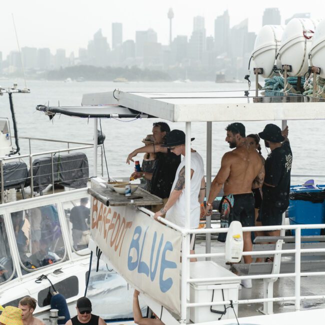 A group of people are gathered on a two-level boat in a harbor. The boat is named "Barefoot Blue." Some individuals are shirtless, and the city skyline is visible in the background under a cloudy sky.