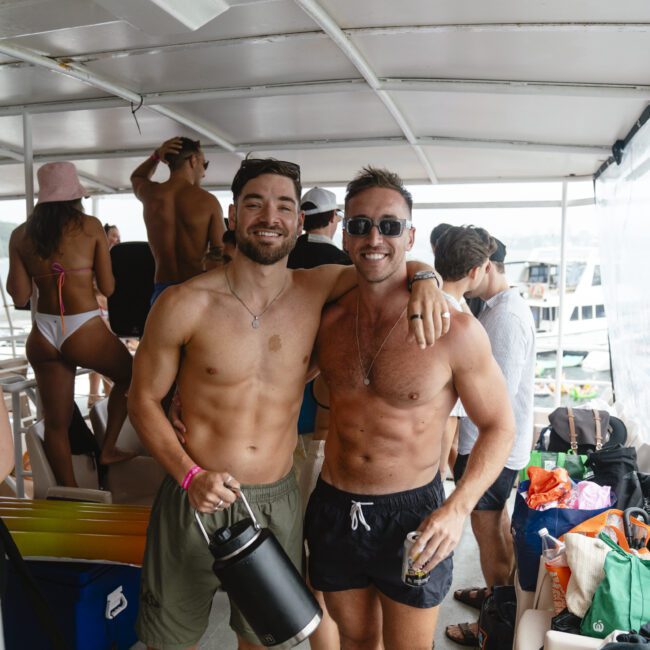 Two shirtless men smiling on a boat. One holds a black jug, and the other wears sunglasses. They're surrounded by other people and various colorful bags and items. The setting appears to be a casual outdoor gathering.