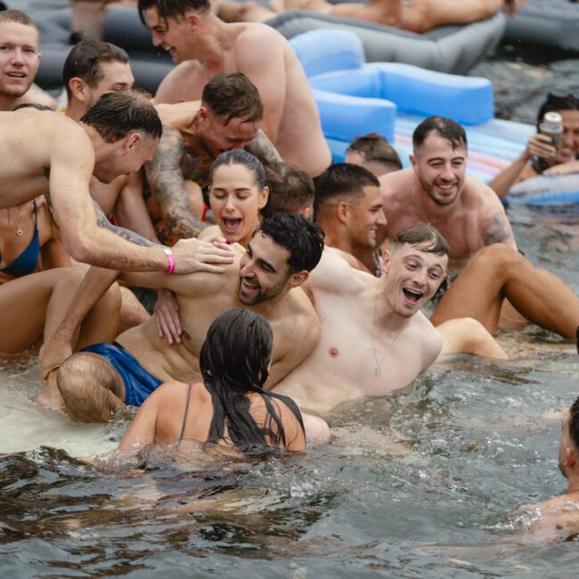 A group of people are enjoying a party in the water, sitting on and around inflatable rafts. They appear happy and are laughing while interacting with each other. Some are drinking beverages, and the scene is lively and festive.