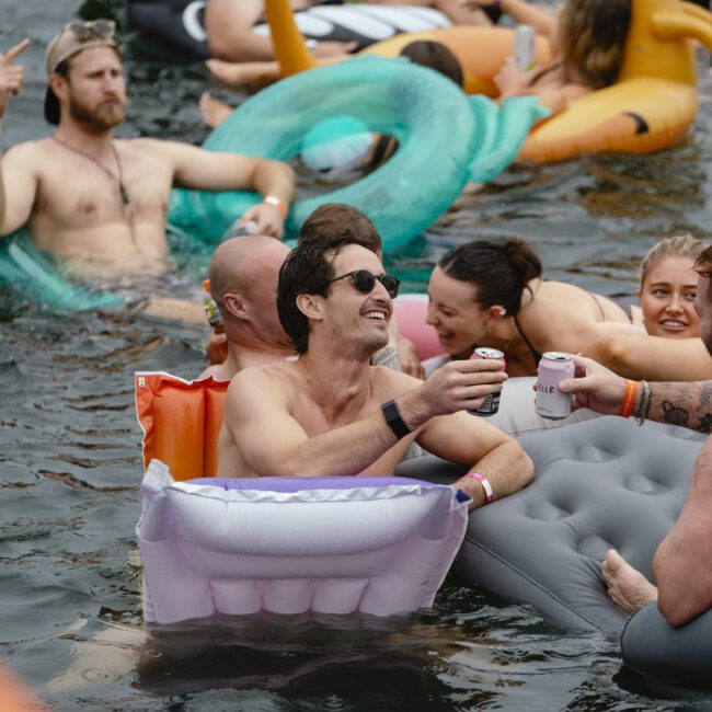 People are enjoying a float on a river with colorful inflatables. A smiling man on a white inflatable holds a canned drink and gestures to others. The group is socializing in the water, surrounded by various floaties and greenery.