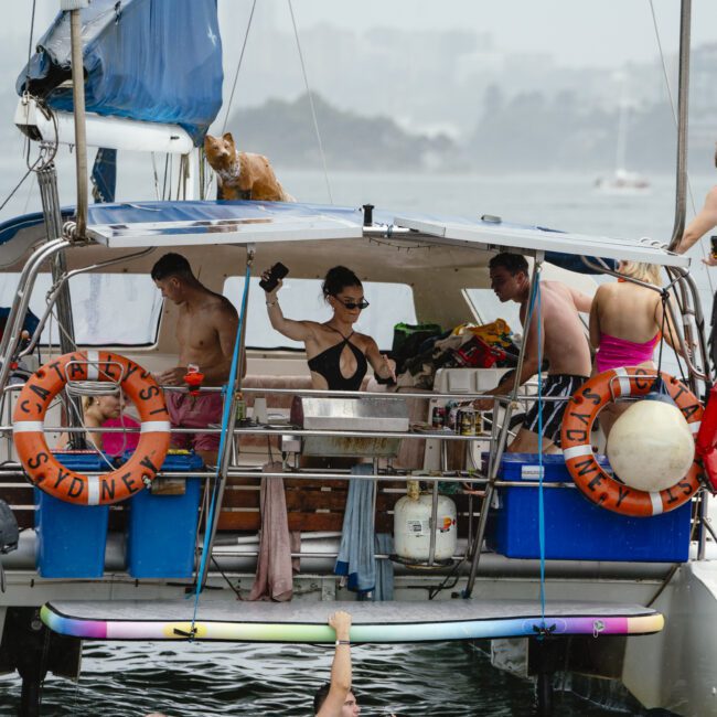A group of people relax on a yacht, with some standing on deck and others swimming in the water nearby. Inflatable rings float around, and a dog is perched on the boat's roof. The backdrop features a hazy view of distant buildings.