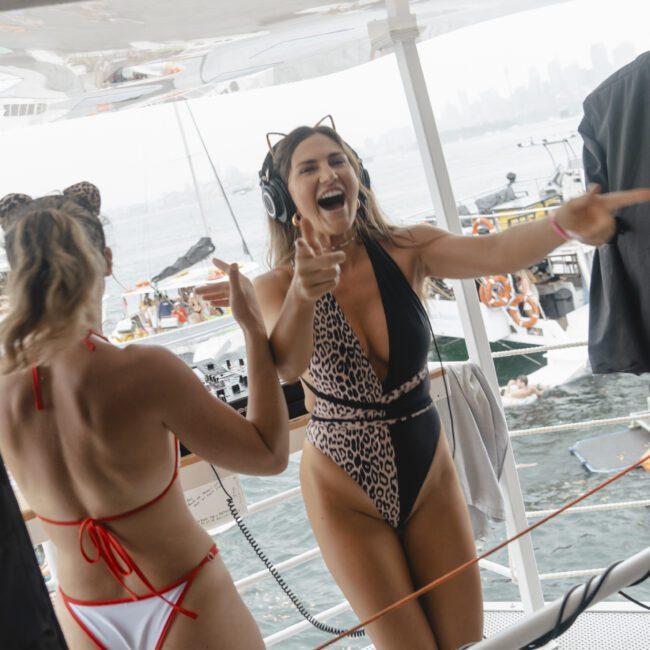 Two women on a boat are wearing animal print swimsuits and DJ headphones, laughing and enjoying music. Boats and water are visible in the background, suggesting a lively outdoor party atmosphere.