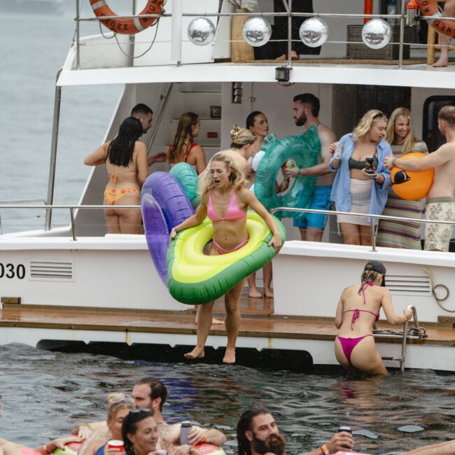 A lively scene on a boat and in the water, with people in swimsuits enjoying a party. A woman with an inflatable ring jumps off the boat. Others float on colorful inflatables in the water. The atmosphere is fun and festive.