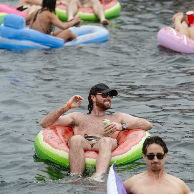 People are floating on colorful inflatable rafts in a lake. A man in a black hat and sunglasses is relaxing on a watermelon-shaped float, holding a drink. Others nearby are enjoying the water and inflatables.