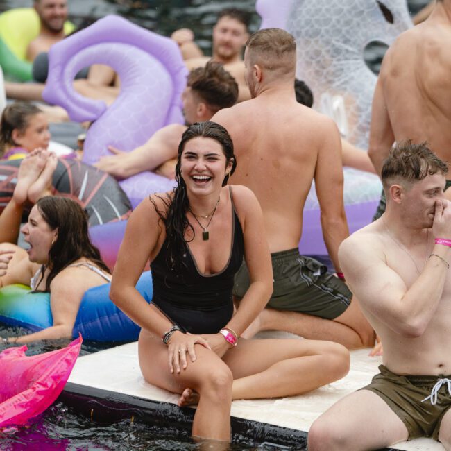 A group of people enjoying a pool party, sitting and standing on inflatable floats. A woman in a black swimsuit laughs, sitting on the pool's edge. The background features colorful floaties and relaxed party attendees.