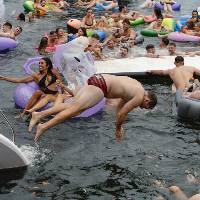 A crowded scene at a lake or beach party with many people enjoying the water. Some are on colorful inflatables, while one person in a swimsuit dives in. Boats and more floaties are visible in the background.
