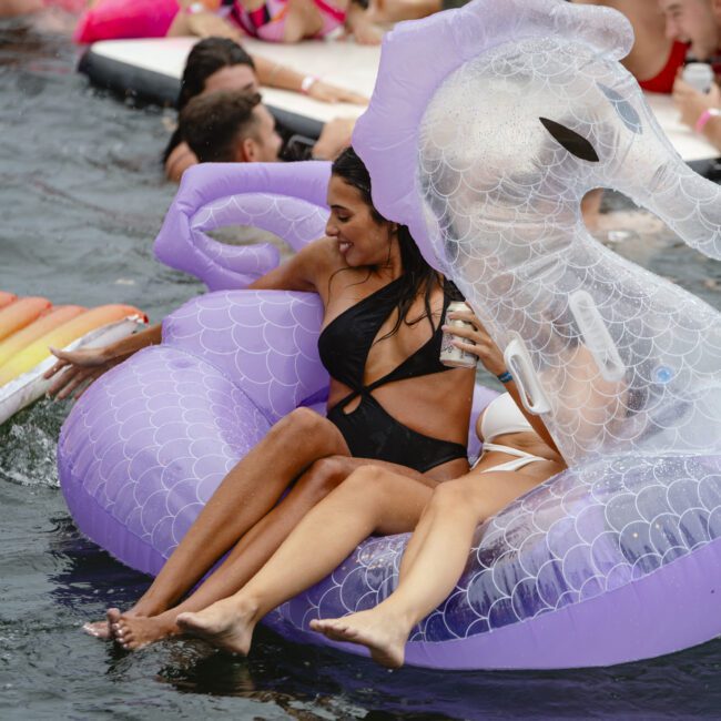 People are having fun at a pool party, with several participants on colorful inflatable floats. A woman in a black swimsuit is on a large purple inflatable swan, surrounded by others enjoying the water.