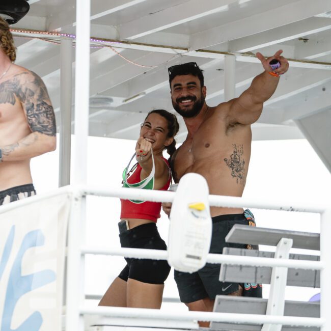 Two people smile and pose playfully on a boat. The man, shirtless, gives a thumbs-up, while the woman points forward. They stand in front of a railing with equipment visible in the background.