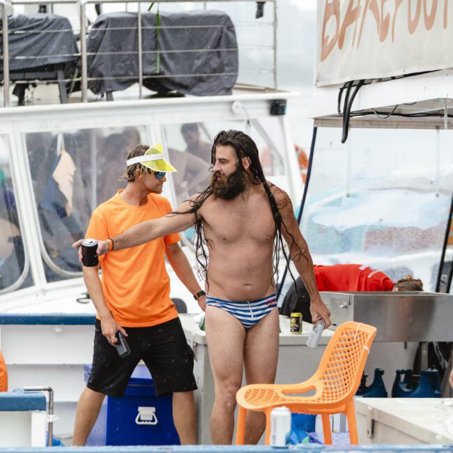 A bearded man with wet hair, wearing striped swim briefs, stands on a boat gesturing with one hand. Another man in an orange shirt and yellow cap stands nearby. An orange chair and signage reading "BAREFOOT" are visible.