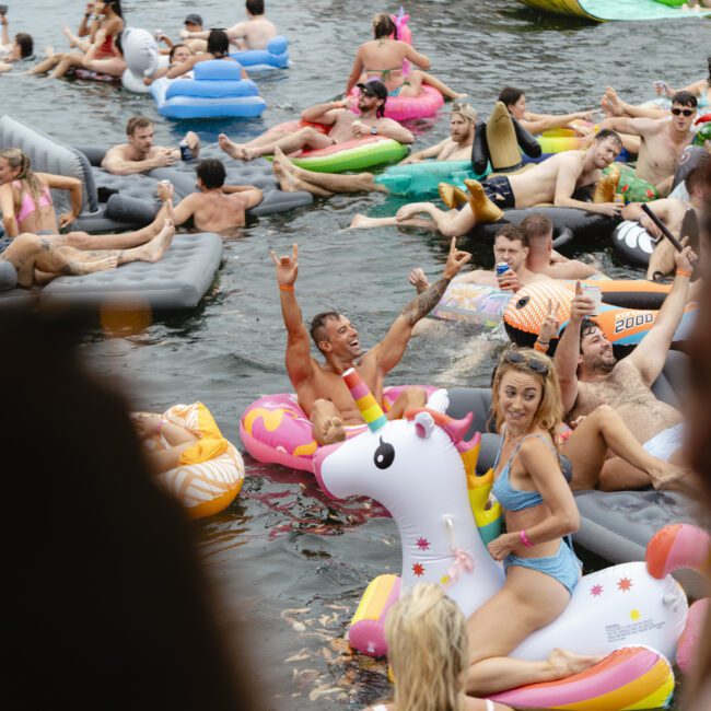 A crowded outdoor scene with people enjoying themselves on inflatable floats in a body of water. A woman in a bikini rides a unicorn float, while others around her relax and have fun on various inflatable shapes.