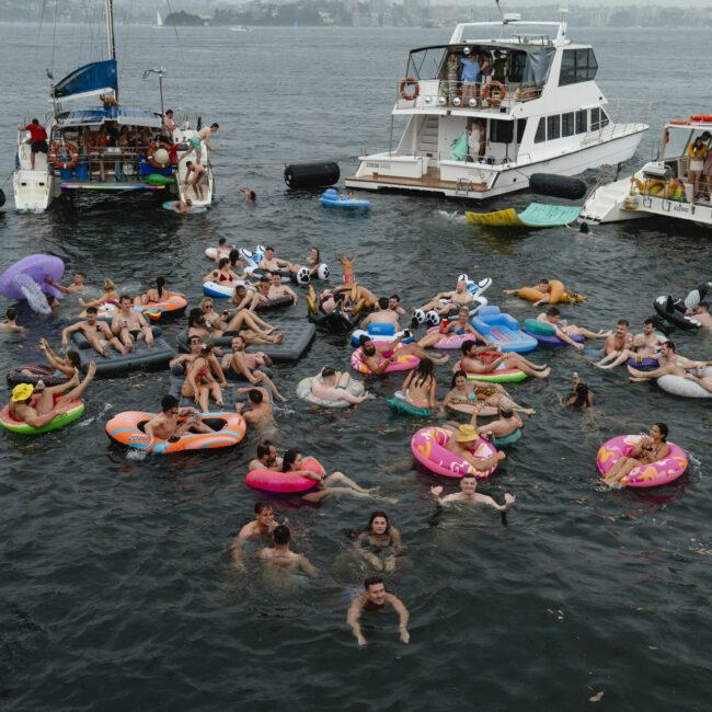 A large group of people in swimsuits float on colorful inflatables in the water near several boats. The scene is lively, with various shaped floats and people swimming, creating a festive atmosphere.