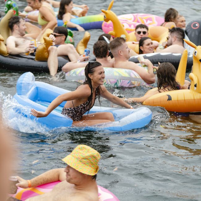 A group of people enjoying a sunny day on a lake, lounging on colorful inflatable rafts shaped like a snail and a blue chair. They seem happy and relaxed, surrounded by water and others also on floaties.