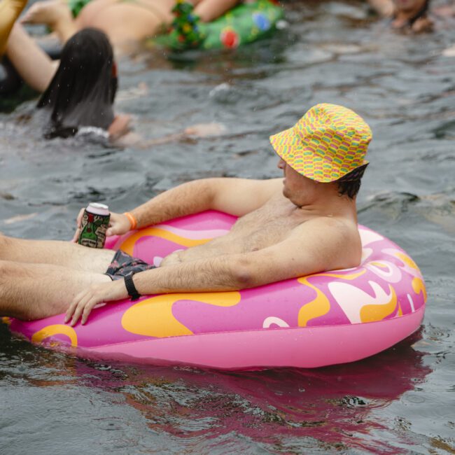 A man relaxes on a pink inflatable ring in the water, wearing a colorful bucket hat and holding a drink. Other people float nearby on different inflatables, enjoying a sunny day on the water.