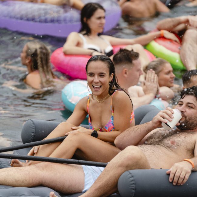 A large group of people enjoying a pool party on inflatable floats. A woman in a colorful swimsuit and a man sipping a drink are in the foreground, looking cheerful. Other partygoers are lounging and socializing in the water.
