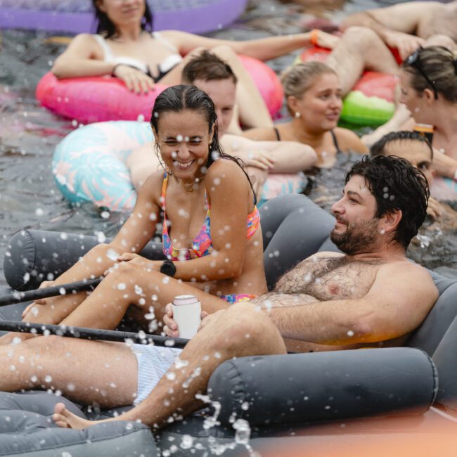 A group of people are relaxing on inflatable floats in a pool. A man and a woman sit together, smiling, as water sprays around them. Other people are gathered in the background, enjoying the water.