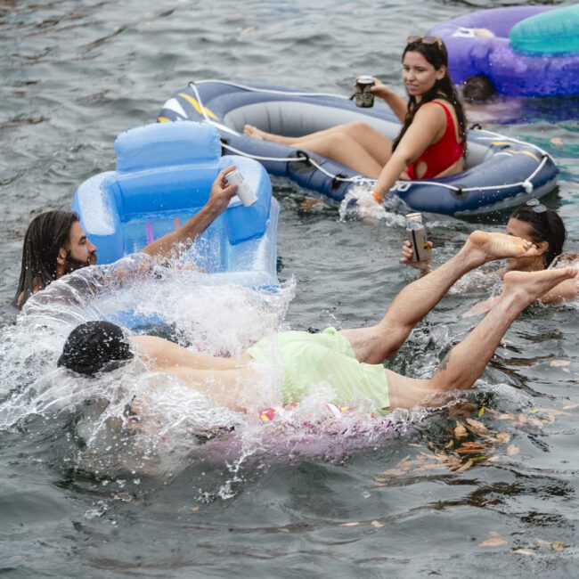 People relax on inflatable loungers in a lake. Two individuals splash into the water, while others float nearby with drinks in hand. The scene is lively and playful against a backdrop of rippling water.