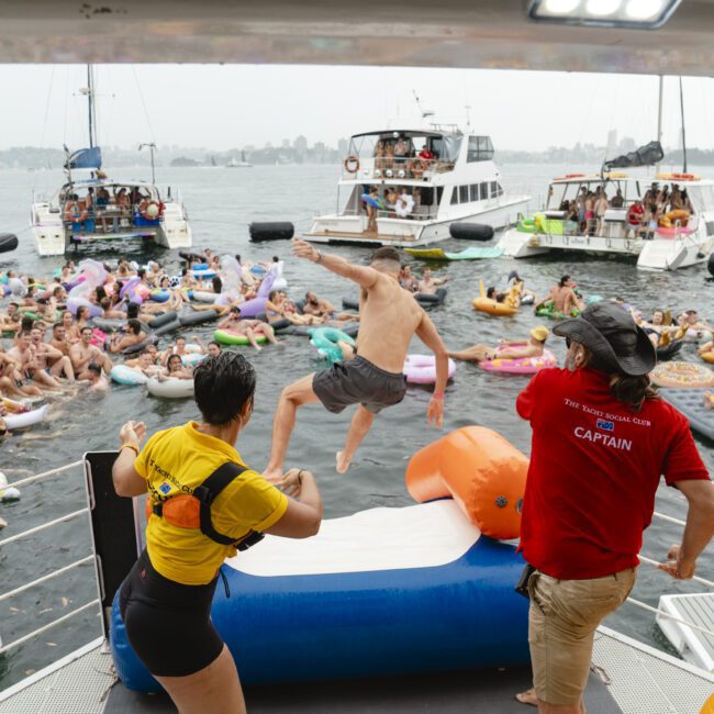 A lively scene of people enjoying a floating party in the water, surrounded by inflatable tubes and nearby boats. A person in mid-air is jumping off an inflatable platform, while others watch and celebrate.