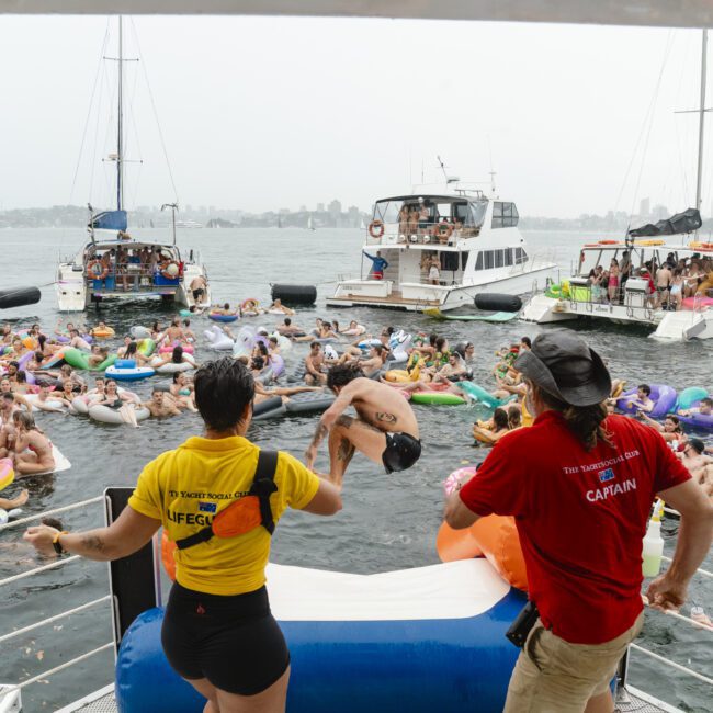 People at a lively boat party, enjoying water activities with numerous inflatables. A person is seen jumping into the water from a boat, surrounded by others watching. Lifeguard and captain are on the boat ensuring safety.