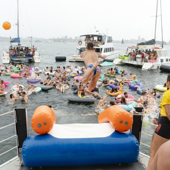 A crowd of people on inflatable pool floats gather in the water near several boats. One person is in mid-air jumping into the water. A lifeguard stands on a platform observing. The scene is lively and festive, suggesting a party or event.