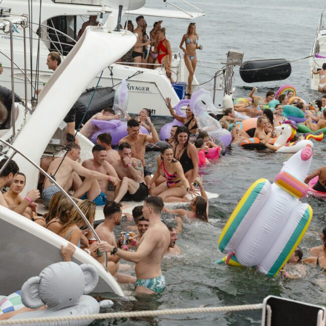 A lively boat party with people enjoying themselves on various inflatable floats in the water. The scene includes a mix of individuals on boats and swimming, surrounded by colorful inflatables.