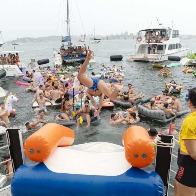 A lively scene on the water with a large group of people on floats and boats. One person is diving into the water, others are sitting on inflatable unicorns and loungers. A lifeguard in a yellow shirt observes the gathering.