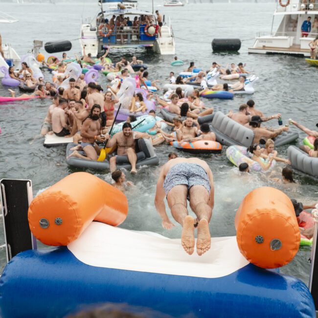 A crowded boat party with people enjoying a summer day on the water. Many are on colorful inflatables, and one person is diving off a slide into the sea. Boats are anchored nearby, and the scene is lively and festive.