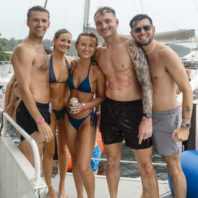 A group of five smiling adults wearing swimsuits stands on a boat. They appear to be enjoying a day out on the water, with life preservers visible in the background. The people are wet, suggesting they may have been swimming.