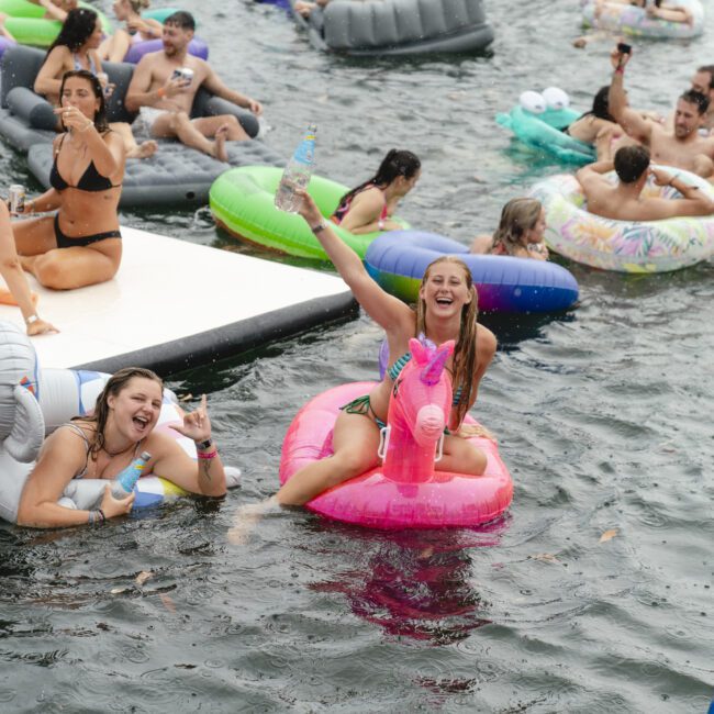 A group of people enjoying a sunny day on a lake, lounging on colorful inflatable rafts. Two women in the foreground, one on a unicorn float and the other on a llama float, smile and hold drinks, surrounded by others in swimsuits.