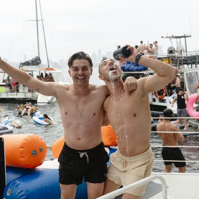 Two shirtless men on a boat having fun; one pouring a drink over himself, laughing. They are surrounded by inflatable tubes on the water, with other boats and people in the background, creating a lively atmosphere.