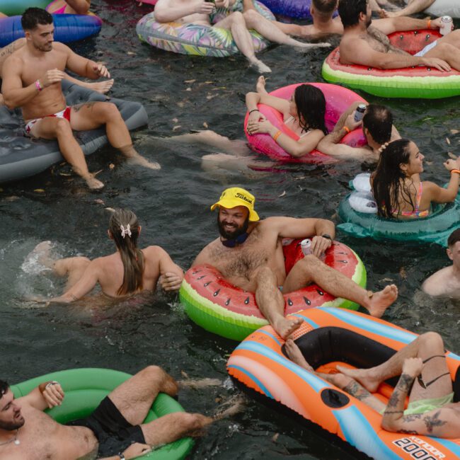 A lively gathering of people enjoying a pool party. They are floating on colorful inflatables in the water, mingling and having fun. The atmosphere is vibrant and festive, with smiles and laughter all around.