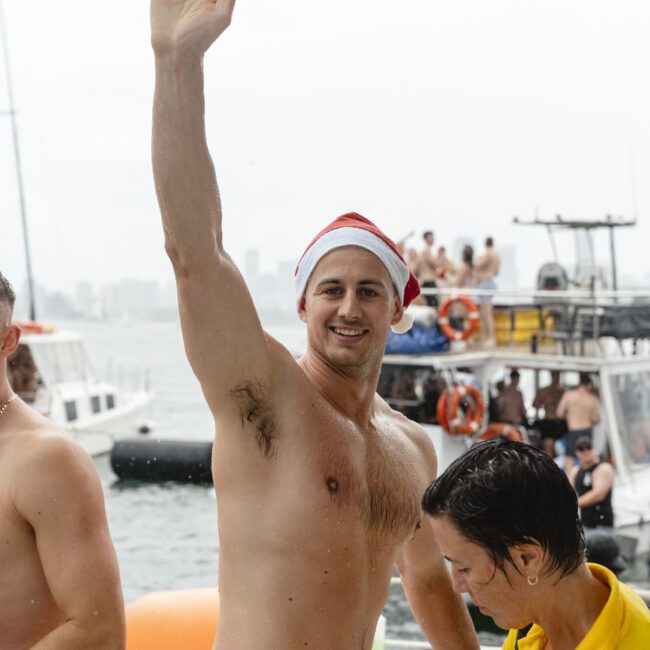 A man wearing a Santa hat and red shorts raises his arm, smiling at a boat party. Another person in a yellow shirt stands nearby. Boats and people are visible in the background on the water.