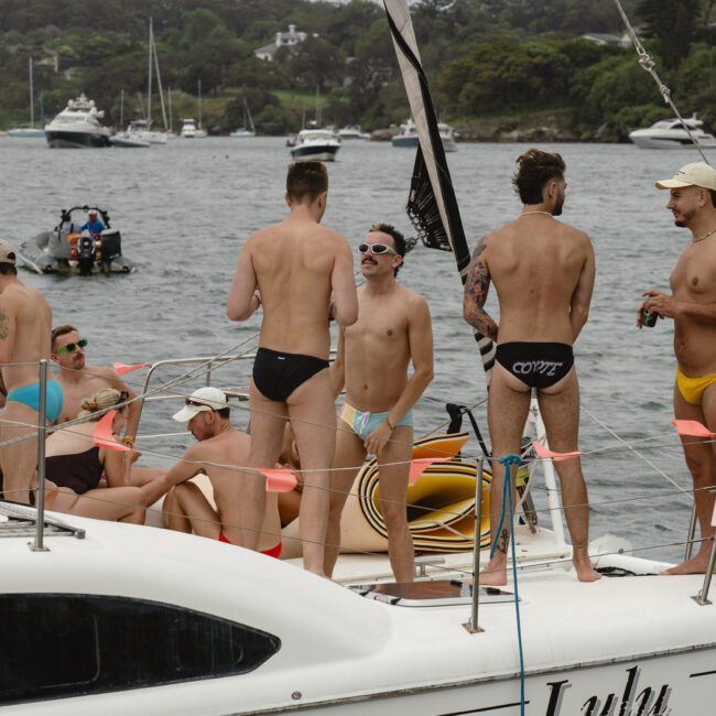 A group of people in swimwear enjoy a sunny day on a yacht. Some are standing and talking, while others sit and relax on the deck. The yacht is on calm water, with other boats and a wooded shoreline in the background.