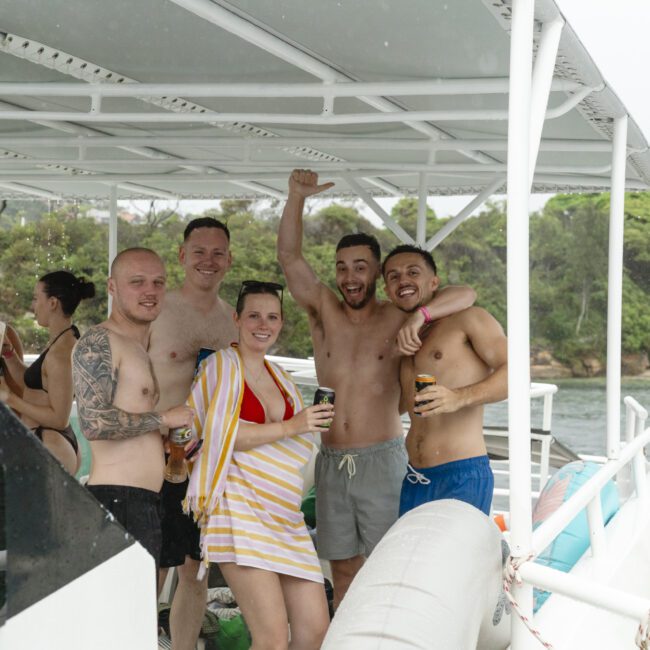 A group of people in swimsuits are standing on a boat, smiling and posing for the camera. They are enjoying a day on the water, with greenery visible in the background. Some hold drinks, and one woman is wearing a striped cover-up.