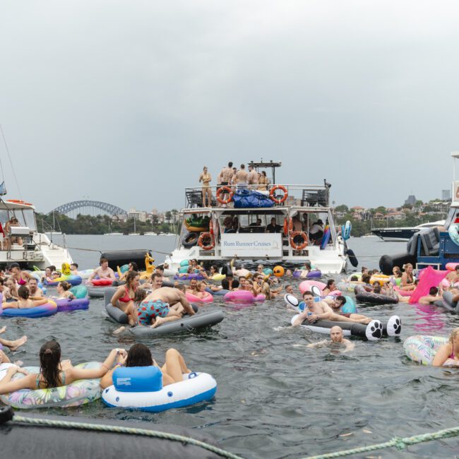 A lively scene of people swimming and lounging on various inflatable floats in the water, surrounded by boats. Some floats are shaped like animals and donuts, while the boats have people observing the festivities. A city skyline is in the background.