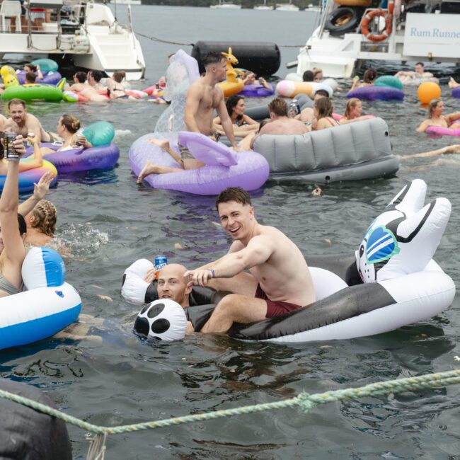 A group of people enjoying a party on a lake, floating on various inflatable rafts and pool toys. Two men are in the foreground, one on an inflatable panda, waving to the camera. Boats and more partygoers are in the background.