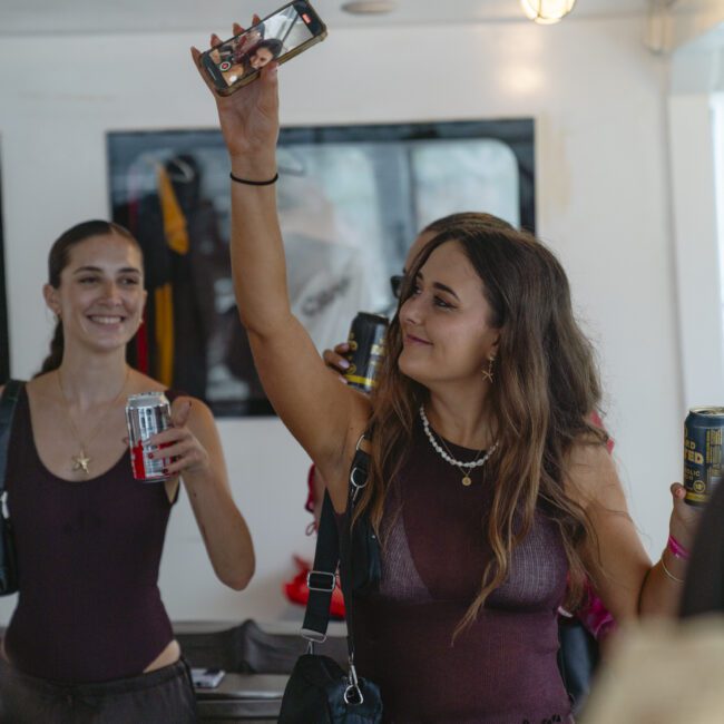 Two women are smiling and holding drinks at an indoor gathering. One woman is raising a phone as if taking a photo or video. Both are wearing dark dresses, with a bar or buffet area in the background.