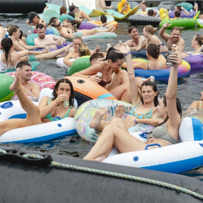 A crowded scene of people enjoying a summer day on a lake, lounging on various inflatable pool floats. The atmosphere is lively with groups chatting, holding drinks, and relaxing in the water.